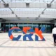 Colorful large letters spelling GRK on a sunny plaza outside a modern glass front building a man with a rolling suitcase walks by