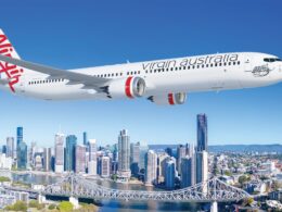 Virgin Australia passenger jet in flight over a city skyline with a river and bridge below against a clear blue sky