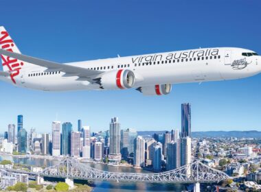 Virgin Australia passenger jet in flight over a city skyline with a river and bridge below against a clear blue sky