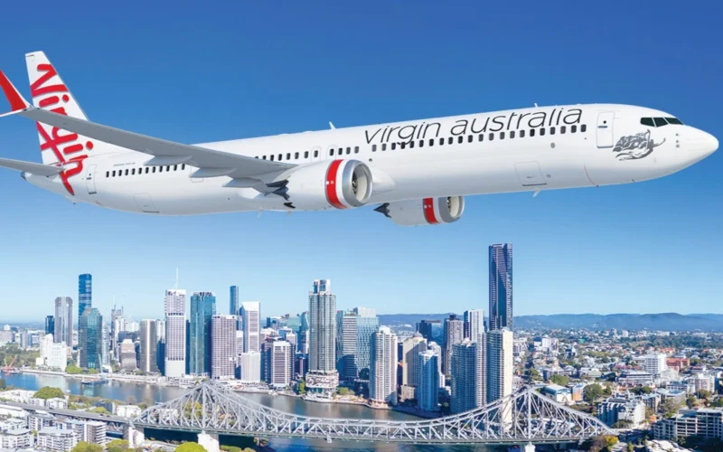 Virgin Australia passenger jet in flight over a city skyline with a river and bridge below against a clear blue sky