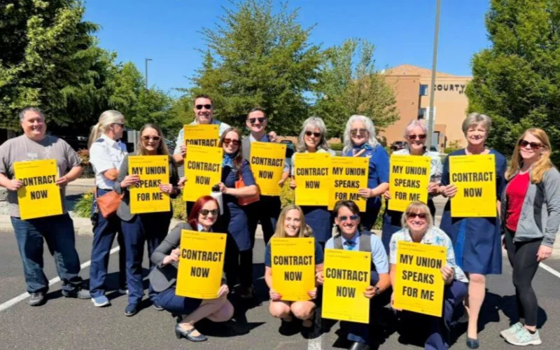 Group of diverse people in a parking lot holding bright yellow signs that read Contract Now and My Union Speaks For Me