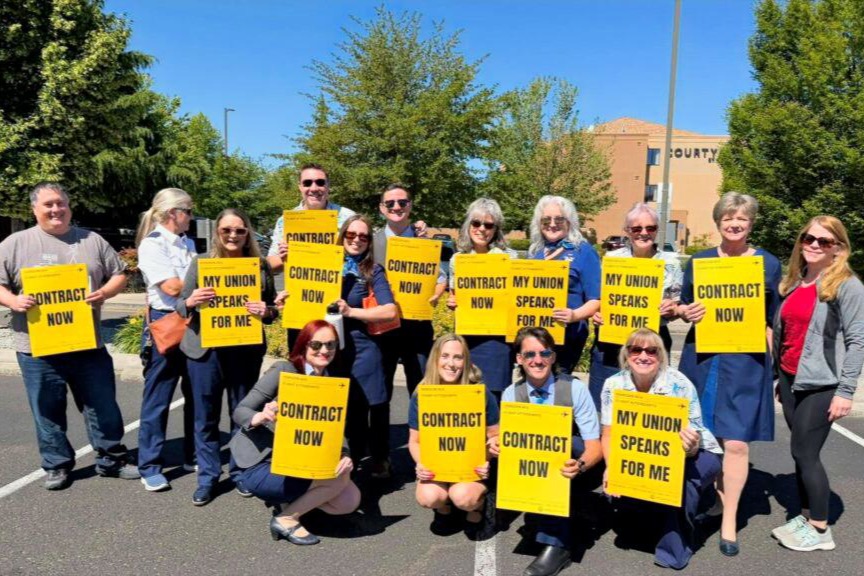 Group of diverse people in a parking lot holding bright yellow signs that read Contract Now and My Union Speaks For Me