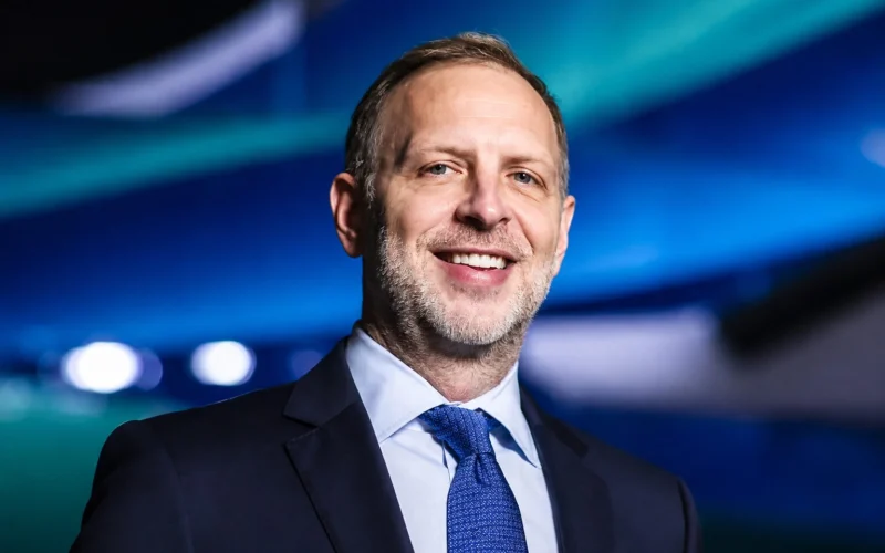 Smiling middle aged man in a dark suit and blue tie with a blue lit blurred background