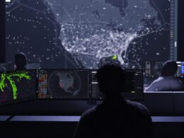 Dark operations room with several monitors displaying a world map of dotted connections a large wall screen shows a dense constellation of white points behind the analysts highlighting monitoring activity
