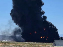 Massive black smoke plume rising from a large fire in an open field flames visible at the base near a low buildingstructure