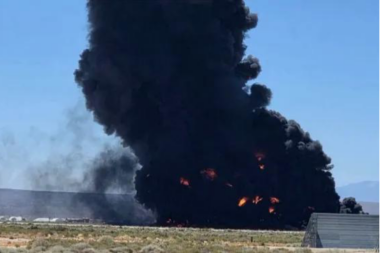Massive black smoke plume rising from a large fire in an open field flames visible at the base near a low buildingstructure