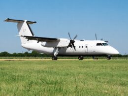 White twinengine turboprop passenger aircraft parked on a grassy airfield with a clear blue sky and a distant tree line