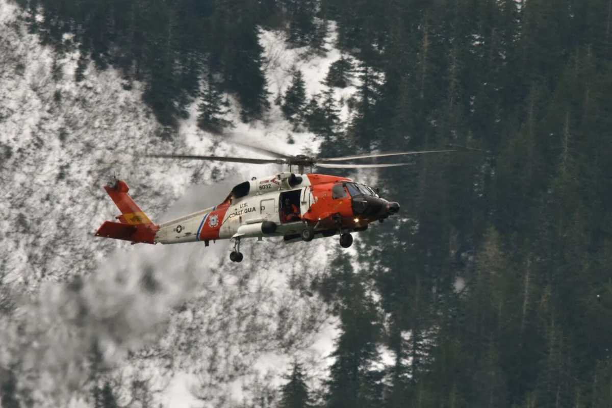 Orange and white US Coast Guard helicopter in flight above a snow covered forest of tall evergreen trees