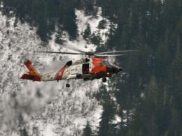 Orange and white US Coast Guard helicopter in flight above a snow covered forest of tall evergreen trees