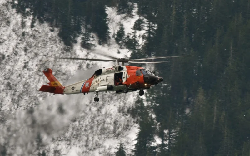 Orange and white US Coast Guard helicopter in flight above a snow covered forest of tall evergreen trees