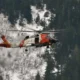 Orange and white US Coast Guard helicopter in flight above a snow covered forest of tall evergreen trees
