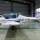 Small white light aircraft with blue accents inside a hangar nose gear down and propeller at left tail marked B 12PP