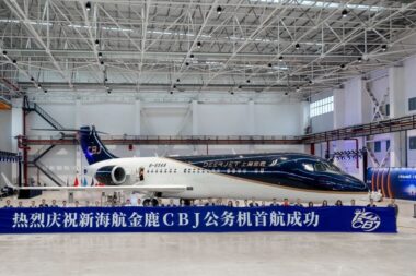 Inside a spacious hangar a dark blue and white private jet sits behind a long blue banner with Chinese text and a row of people posing in front