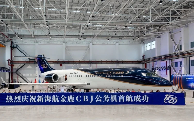 Inside a spacious hangar a dark blue and white private jet sits behind a long blue banner with Chinese text and a row of people posing in front