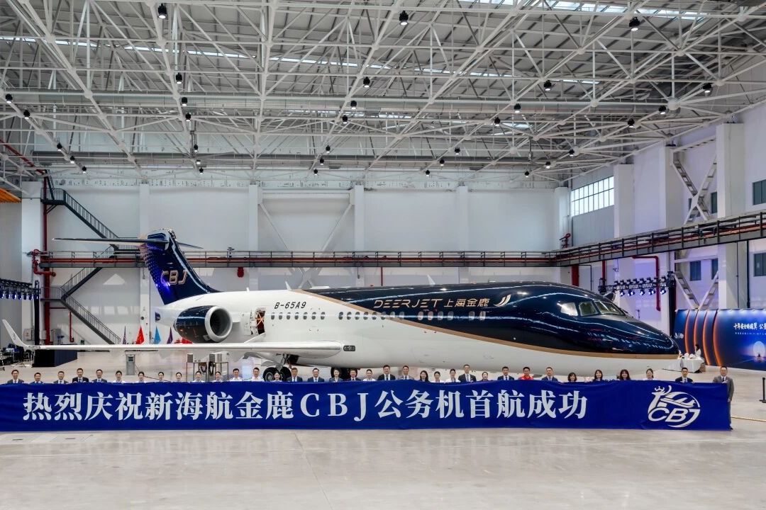 Inside a spacious hangar a dark blue and white private jet sits behind a long blue banner with Chinese text and a row of people posing in front