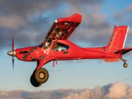 Red aerobatic airplane in midflight propeller blurred blue sky with clouds in the background showcasing its branding on the fuselage