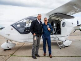 Two men pose smiling beside a small white private turboprop aircraft with the door open on a runway apron