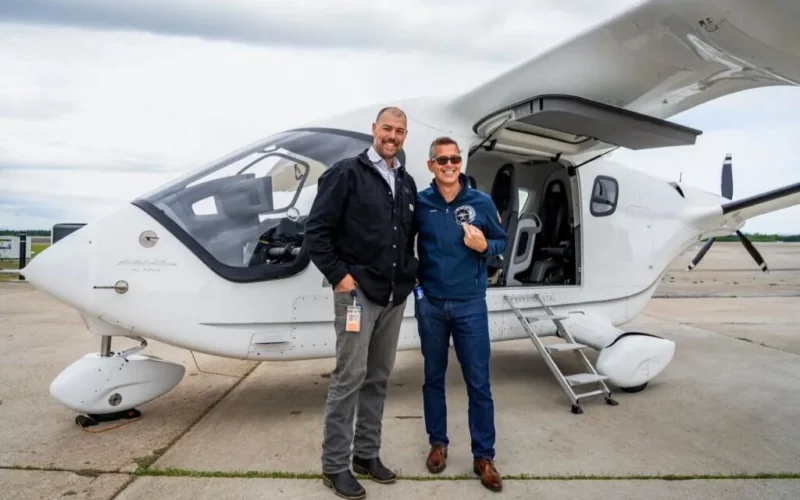 Two men pose smiling beside a small white private turboprop aircraft with the door open on a runway apron