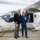 Two men pose smiling beside a small white private turboprop aircraft with the door open on a runway apron