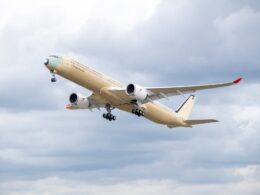 Beige passenger jet with red wingtip climbs into a cloudy sky during takeoff landing gear down