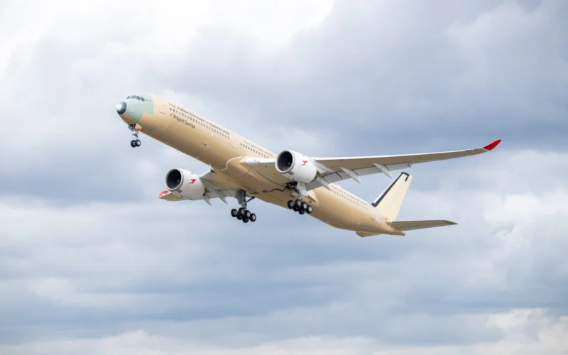 Beige passenger jet with red wingtip climbs into a cloudy sky during takeoff landing gear down