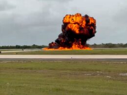 Large explosion on a runway with a massive orange flame plume and dark smoke rising into the sky surrounded by grass fields