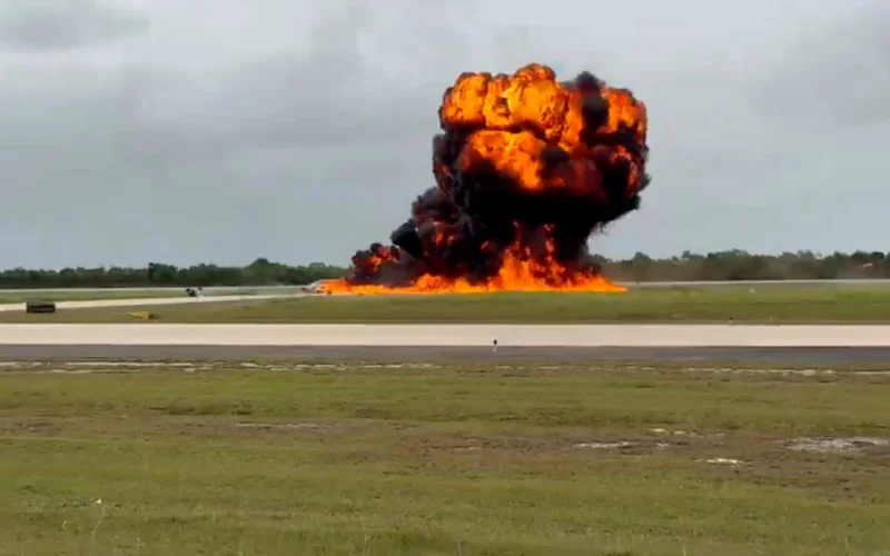 Large explosion on a runway with a massive orange flame plume and dark smoke rising into the sky surrounded by grass fields