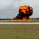 Large explosion on a runway with a massive orange flame plume and dark smoke rising into the sky surrounded by grass fields