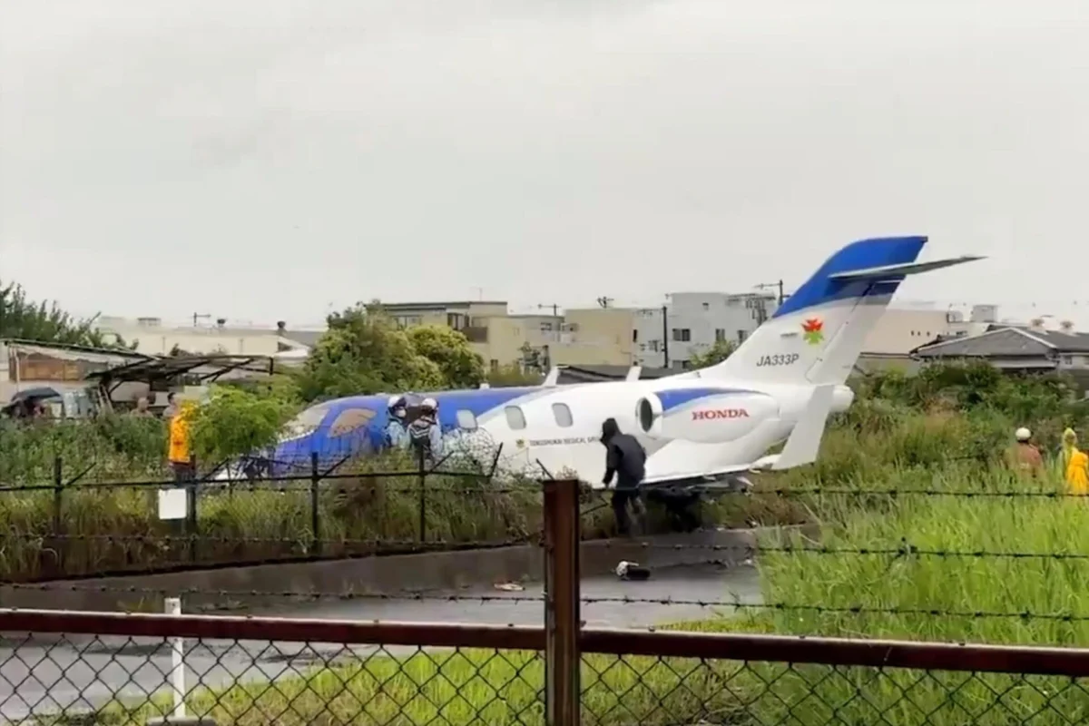 A small white and blue jet fuselage with a blue tail parked on a grassy area near a fence people in reflective vests stand nearby suggesting an incident or recovery scene