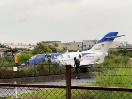A small white and blue jet fuselage with a blue tail parked on a grassy area near a fence people in reflective vests stand nearby suggesting an incident or recovery scene