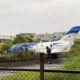 A small white and blue jet fuselage with a blue tail parked on a grassy area near a fence people in reflective vests stand nearby suggesting an incident or recovery scene