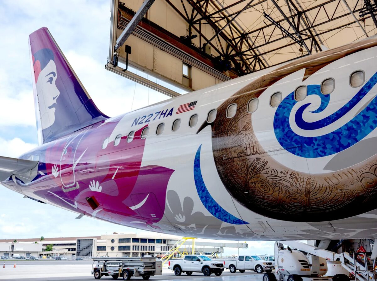 image Tail of a colorful airplane with purplepink design US flag and registration N227HA parked at an airport with ground support equipment nearby
