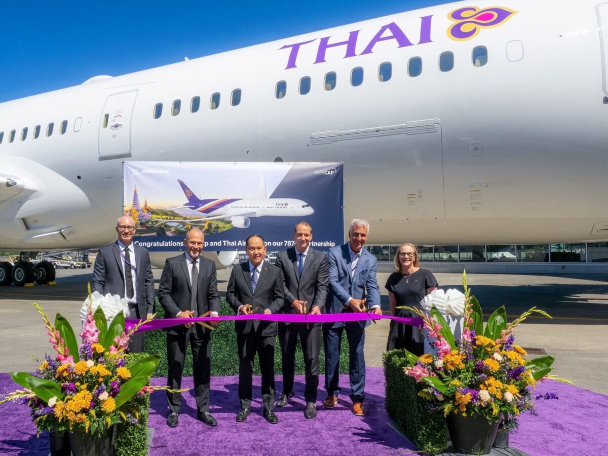 Group of executives cutting a purple ribbon in front of a Thai Airways plane during a ceremony with colorful flower arrangements nearby