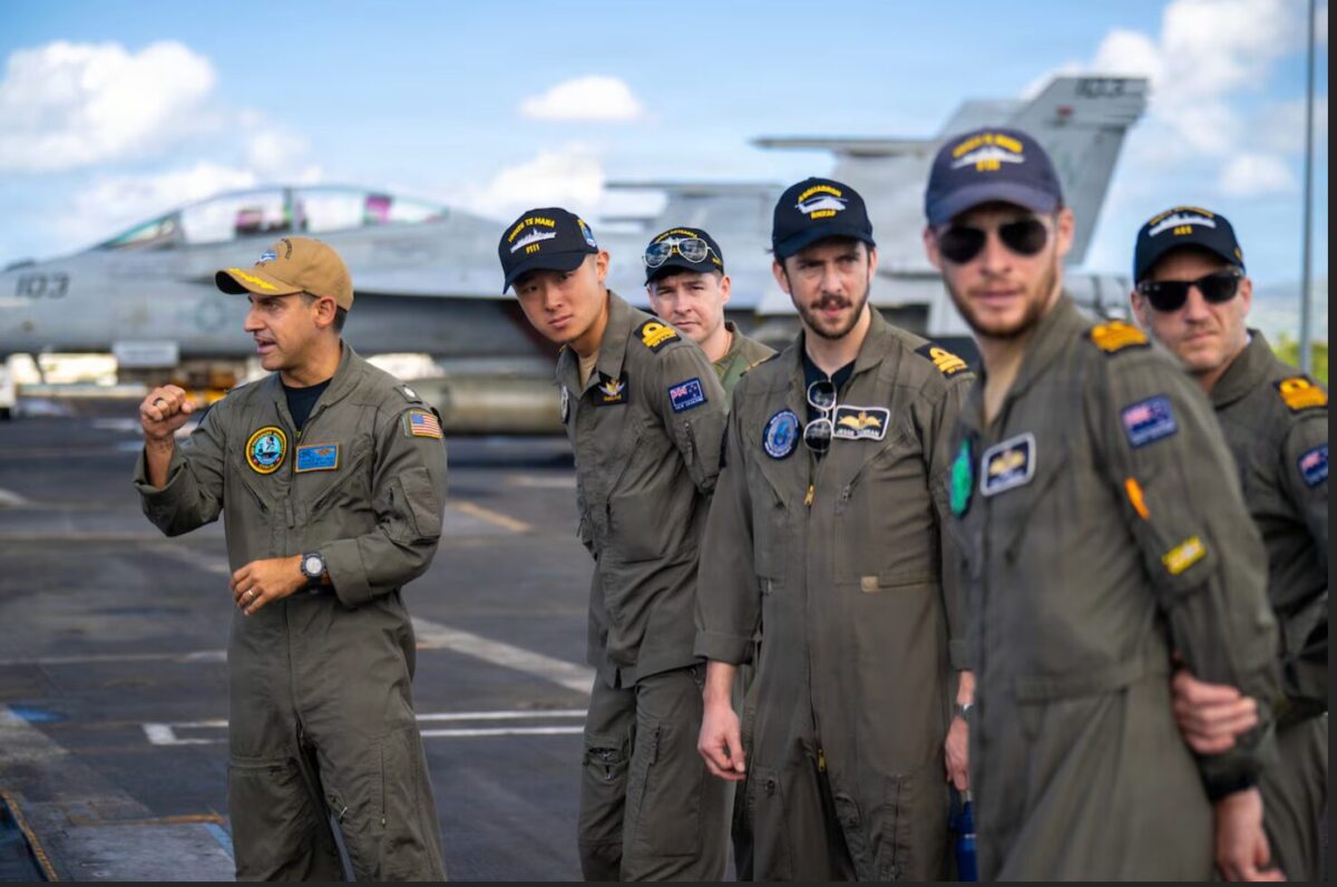 image Group of pilots in olive flight suits posing on an aircraft carrier deck with fighter jets in the background blue sky above