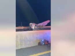 Night scene of a crashed small airplane on a highway barrier with firefighters examining the wreckage and debris on the roadlighted by emergency vehicles