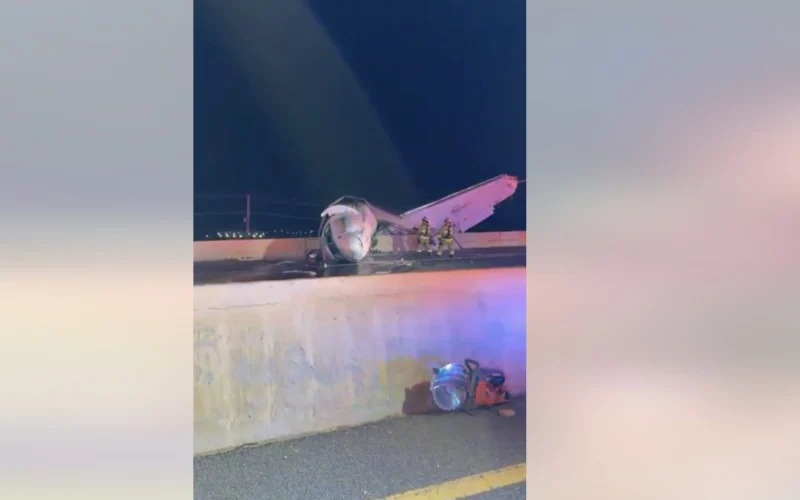 Night scene of a crashed small airplane on a highway barrier with firefighters examining the wreckage and debris on the roadlighted by emergency vehicles
