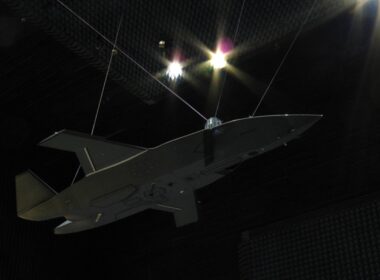 Unmanned aerial vehicle, gray flying-wing drone, suspended by cables inside an anechoic test chamber at a sound-testing facility.