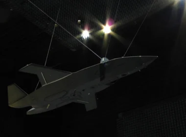 Unmanned aerial vehicle, gray flying-wing drone, suspended by cables inside an anechoic test chamber at a sound-testing facility.