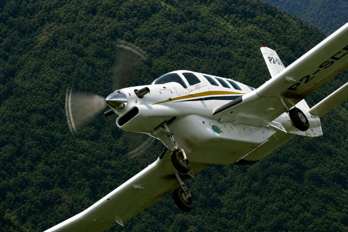 pac75032 White single engine airplane with a gold stripe banking in the air above a forested hillside