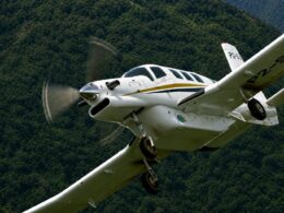 White single engine airplane with a gold stripe banking in the air above a forested hillside