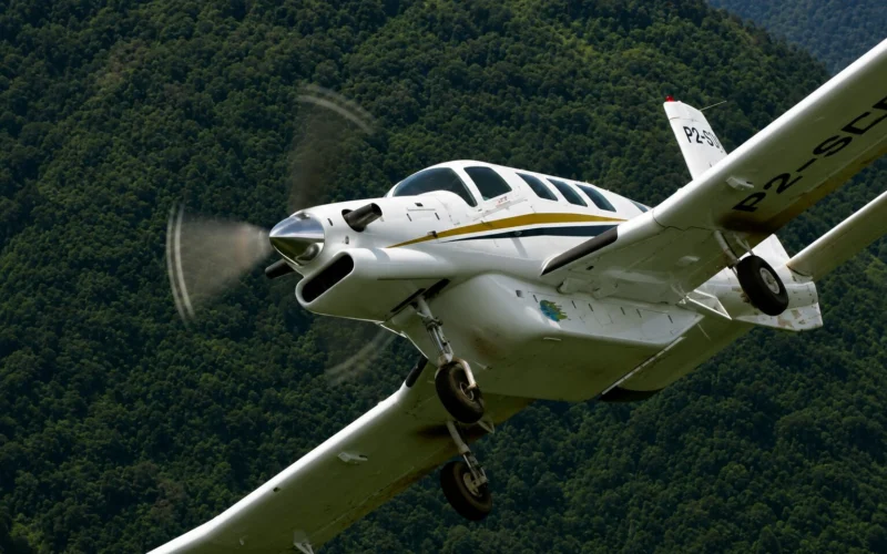 White single engine airplane with a gold stripe banking in the air above a forested hillside