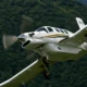 White single engine airplane with a gold stripe banking in the air above a forested hillside
