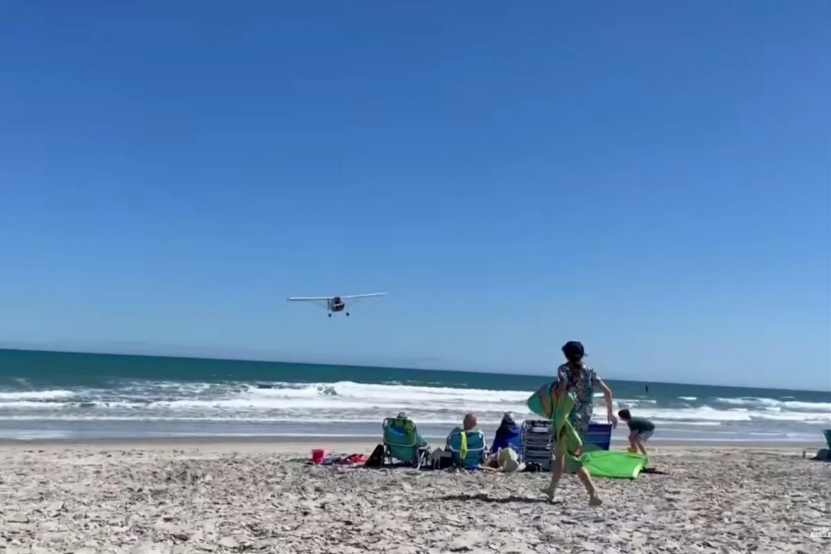 planeoverbeach Small airplane flying low over a sunny beach as people relax on beach chairs and carry surfboards near the shore