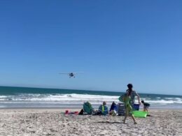 Small airplane flying low over a sunny beach as people relax on beach chairs and carry surfboards near the shore