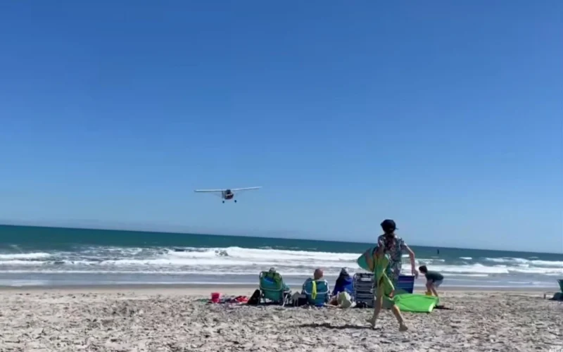 Small airplane flying low over a sunny beach as people relax on beach chairs and carry surfboards near the shore