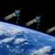 Three blue satellite like structures with extended robotic arms float above Earths curved horizon in space against black sky