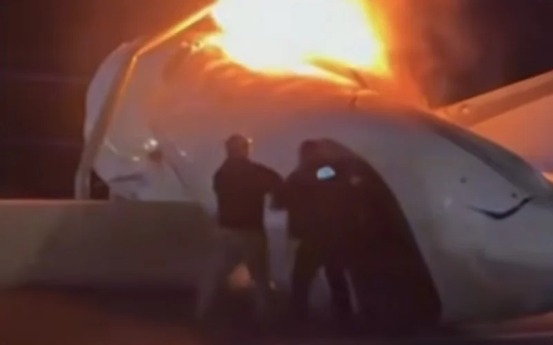 Airplane fuselage on fire with bright flames silhouettes of responders at a nighttime crash site