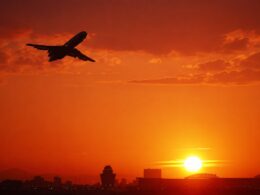 Airplane silhouette flying left to right over a city skyline at a fiery orange sunset