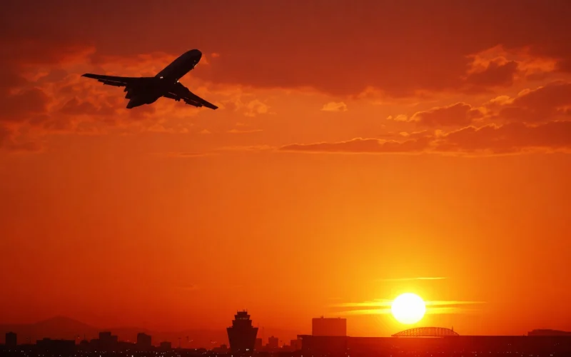 Airplane silhouette flying left to right over a city skyline at a fiery orange sunset