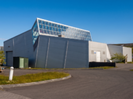 Modern industrial building with sloped gray walls and a glass roofed upper section against a blue sky